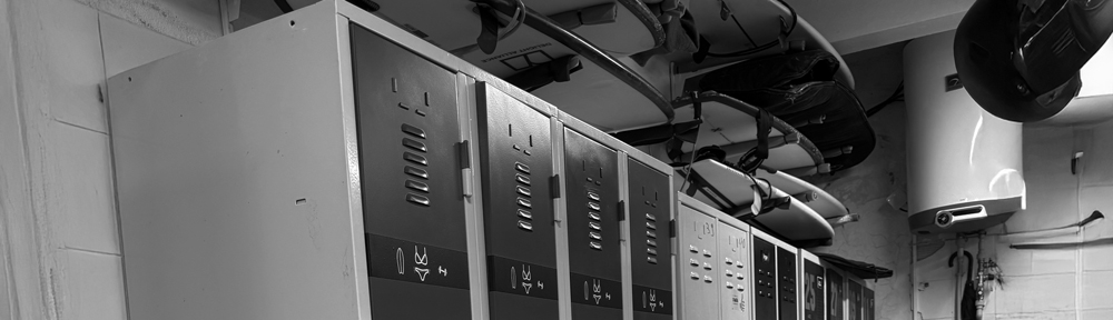 Shaper Liam Bulgen stojí ve své dílně, průhled na tenisové hřiště, surfboard na stole 