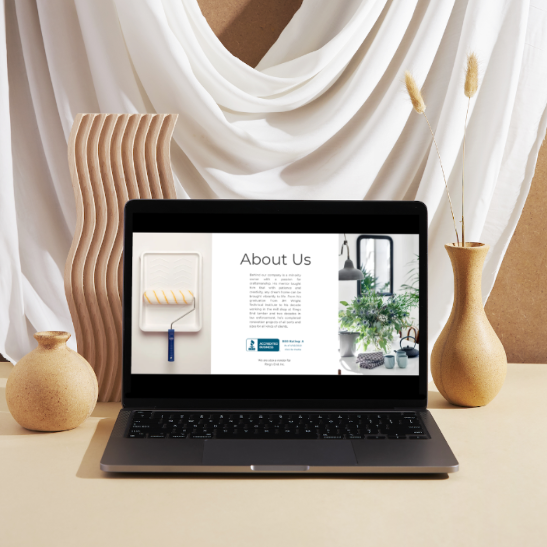 Laptop on a table displaying an About Us page with a paint roller and a plant image, surrounded by ceramic vases and a wavy wooden decor against white draped fabric.