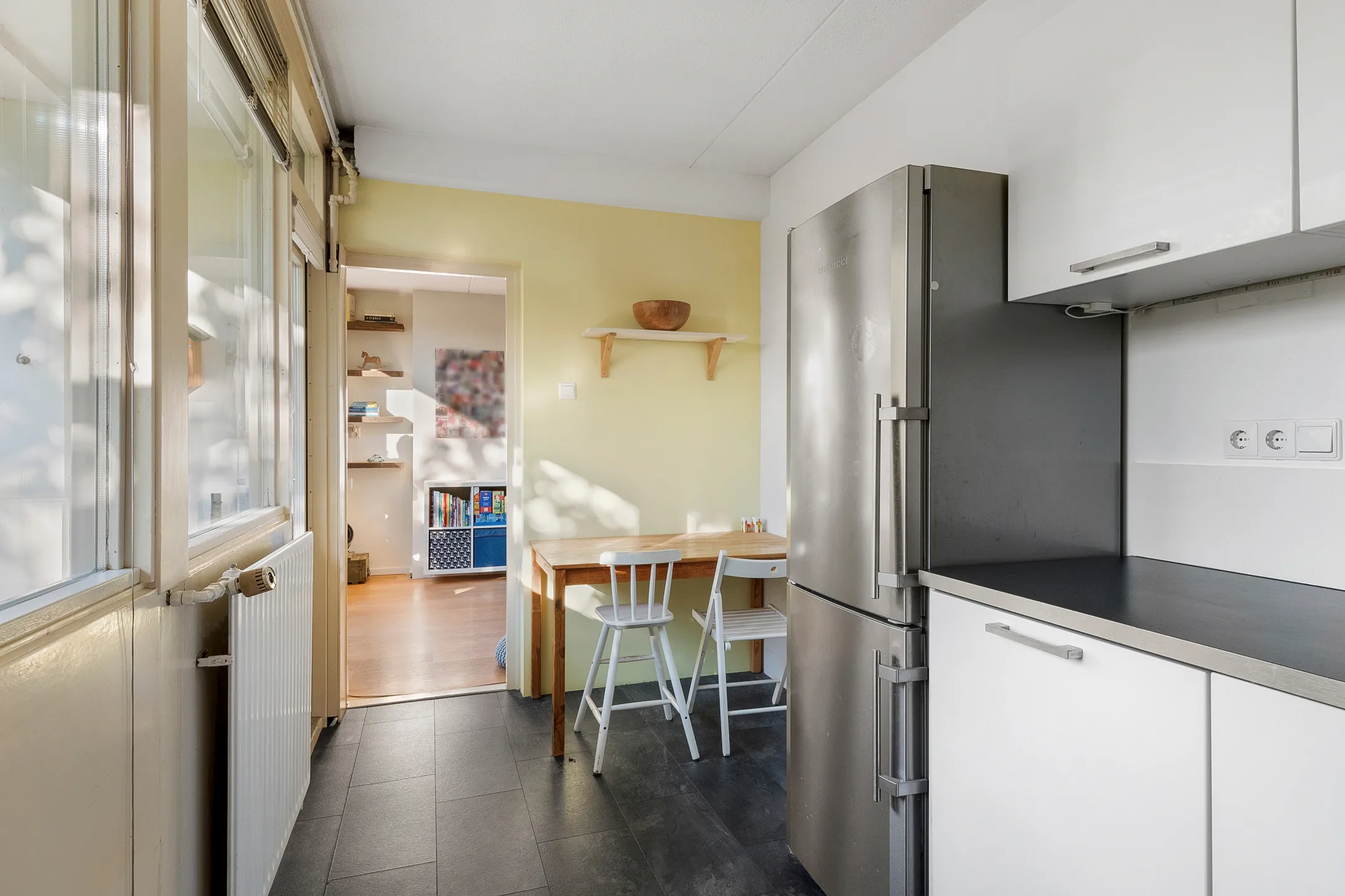 Keuken met witte kasten, een grote koelkast, houten tafel en twee witte stoelen tegen een gele muur.