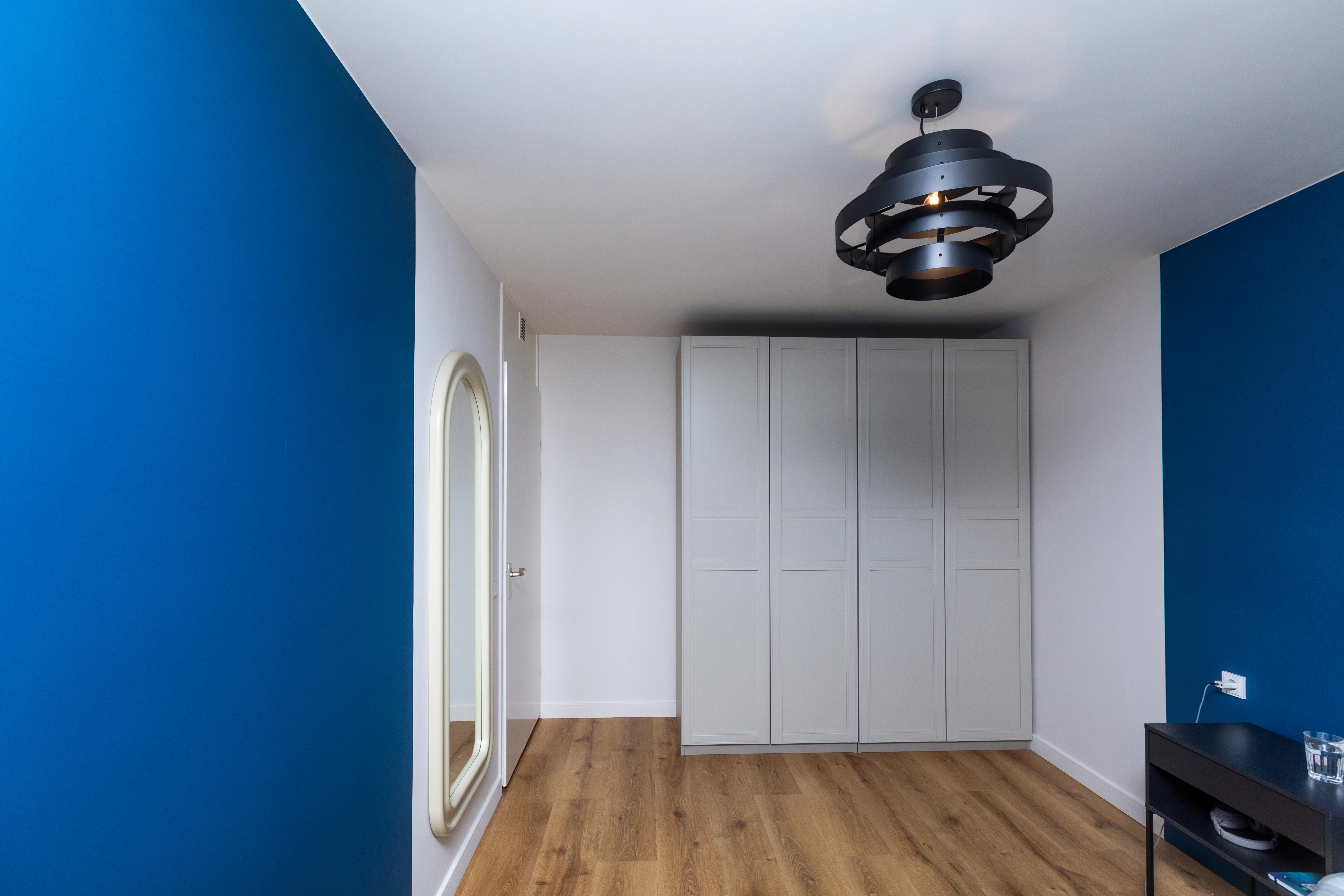 Slaapkamer met blauw geschilderde muren, grote spiegel, grijze kast en houten vloer.