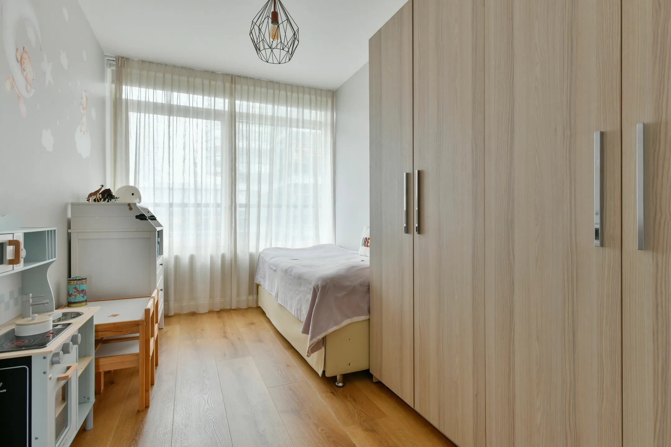 Rustige kinderkamer met een bed, houten kast, speelhoek en zachte gordijnen voor het raam.