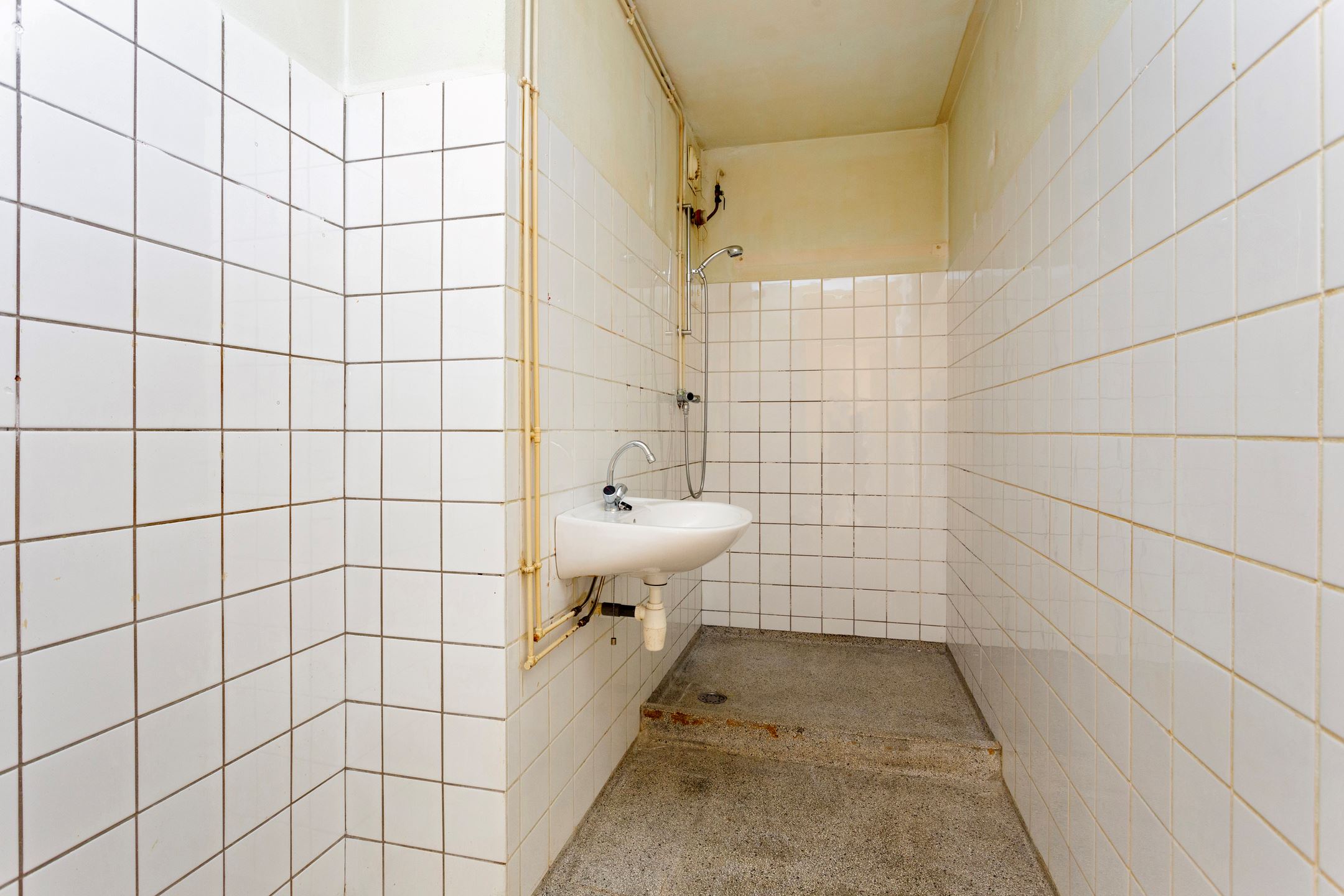 Kleine doucheruimte met witte tegels, een wastafel aan de muur en een douchekop erboven.
