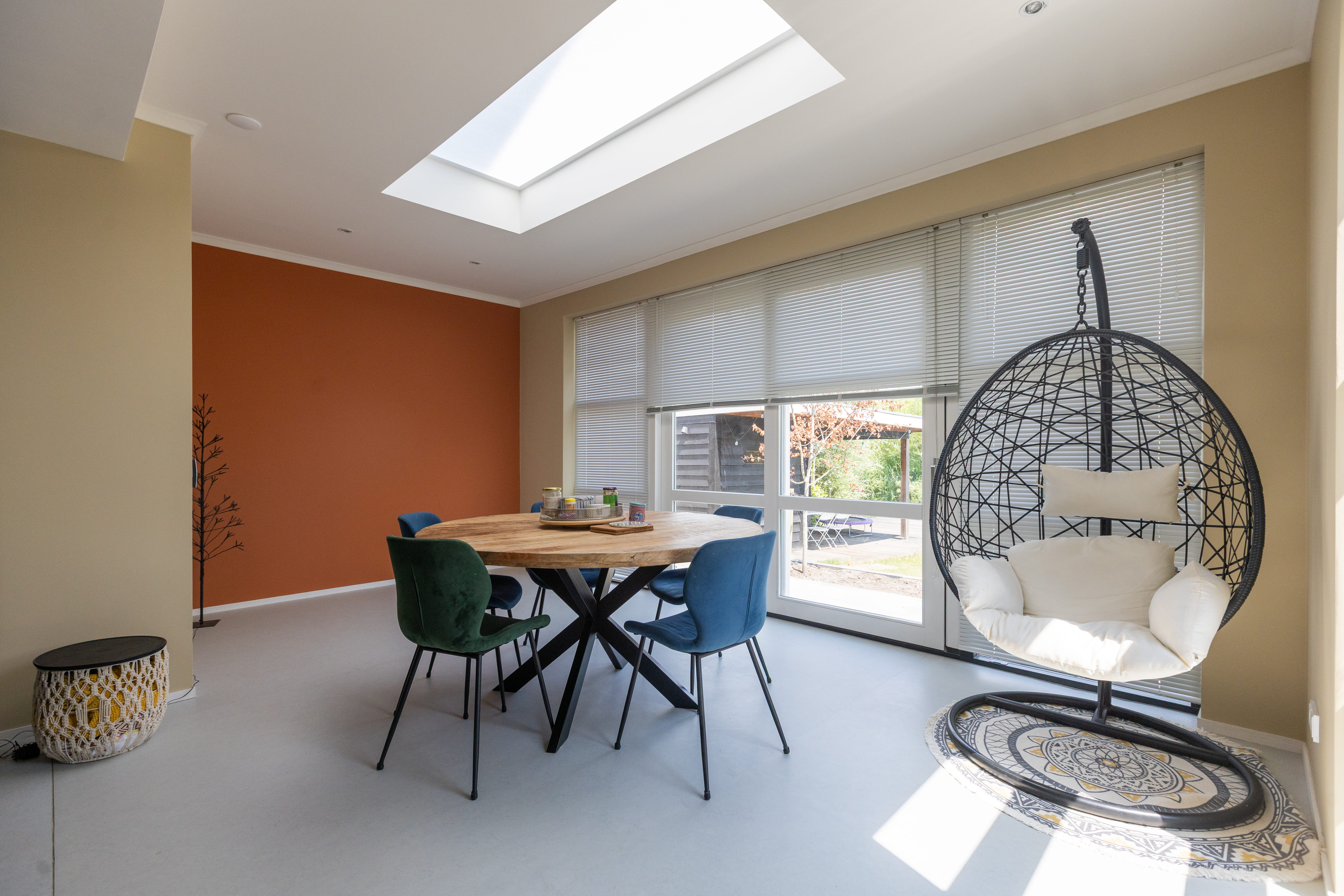 Eetkamer met ronde houten tafel, verschillende gekleurde stoelen en hangstoel bij het raam met luiken.