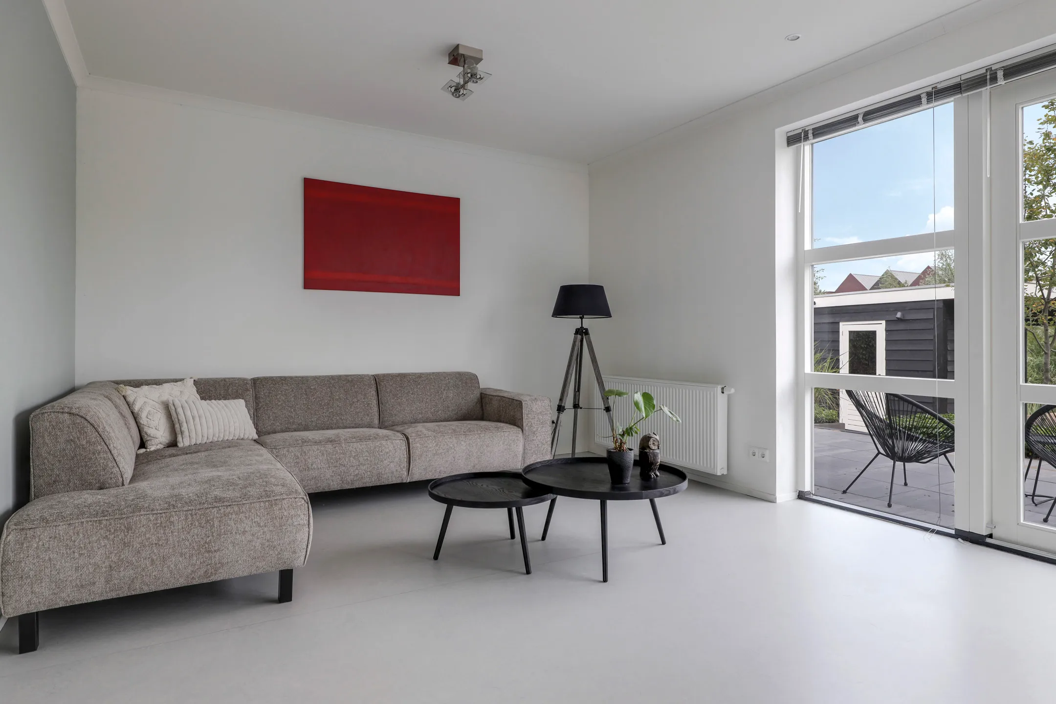 Modern woonkamer met een grijze hoekbank, twee zwarte tafels, vloerlamp en groot raam naar terras.