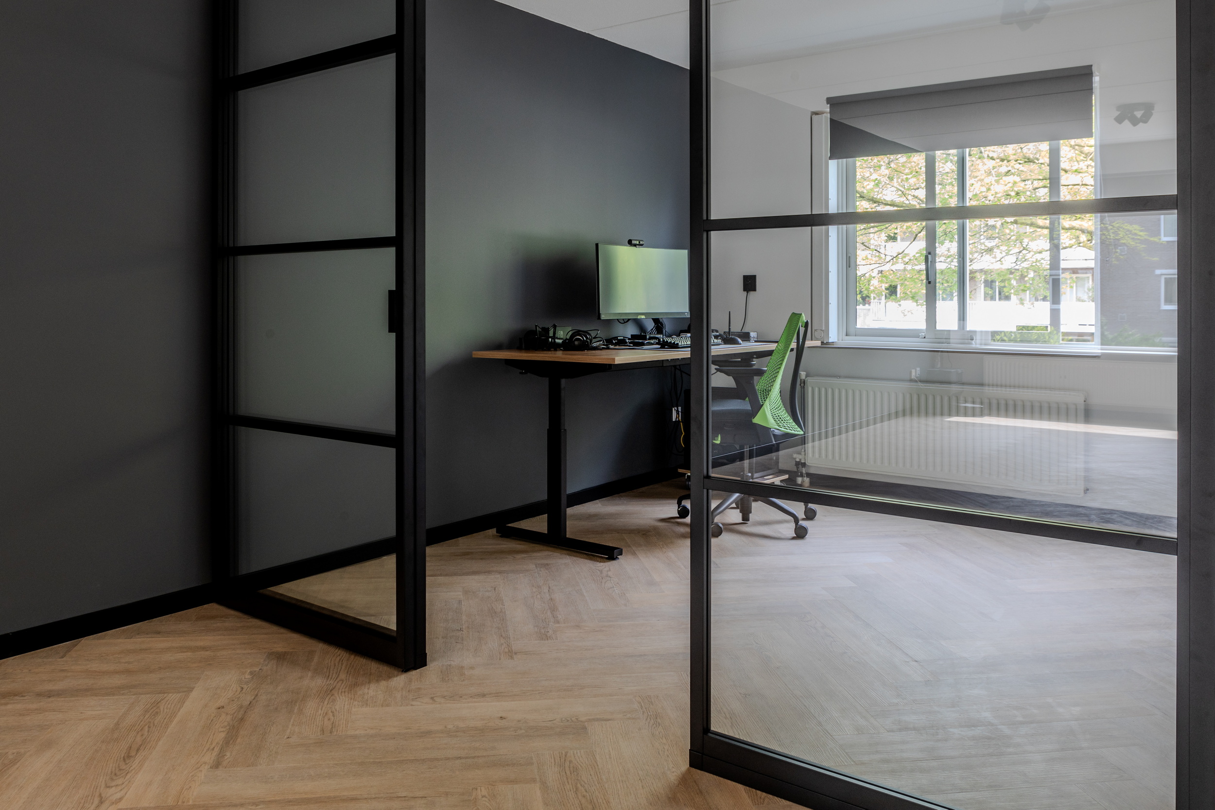 Modern kantoorruimte met glazen wanden, bureau met computer en groene bureaustoel bij raam.