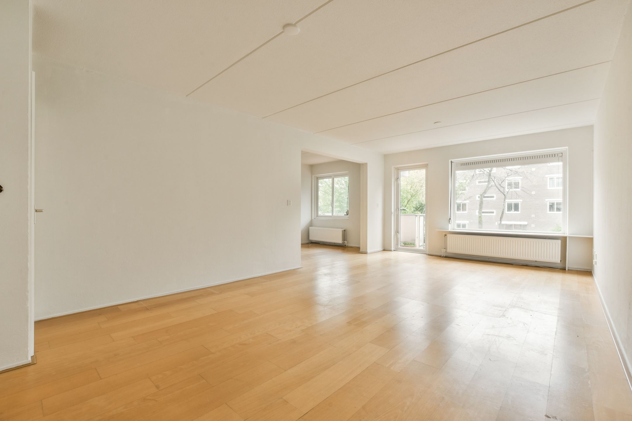 Lege woonkamer met licht houten vloer, grote ramen en witte muren in een appartement.