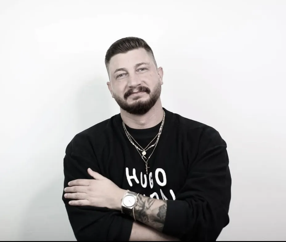 Mann mit kurzem Haar und Bart trägt schwarzen Pullover mit Aufschrift, Halsketten und Armbanduhr, verschränkt die Arme und lächelt.