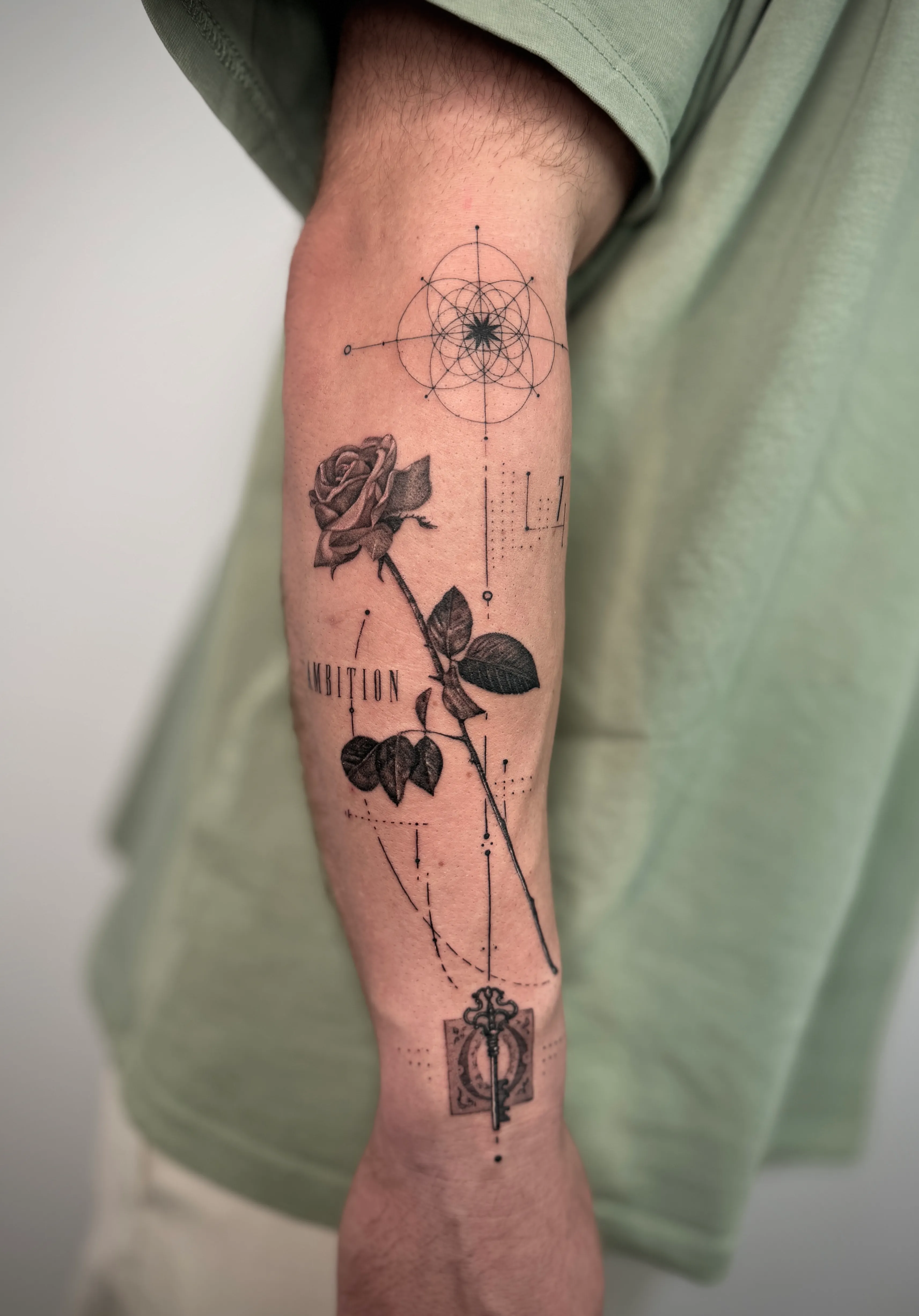 Schwarz-weiße Tätowierung am Unterarm mit einer Rose, einem Schlüssel und geometrischen Linien mit dem Wort ‚AMBITION‘.