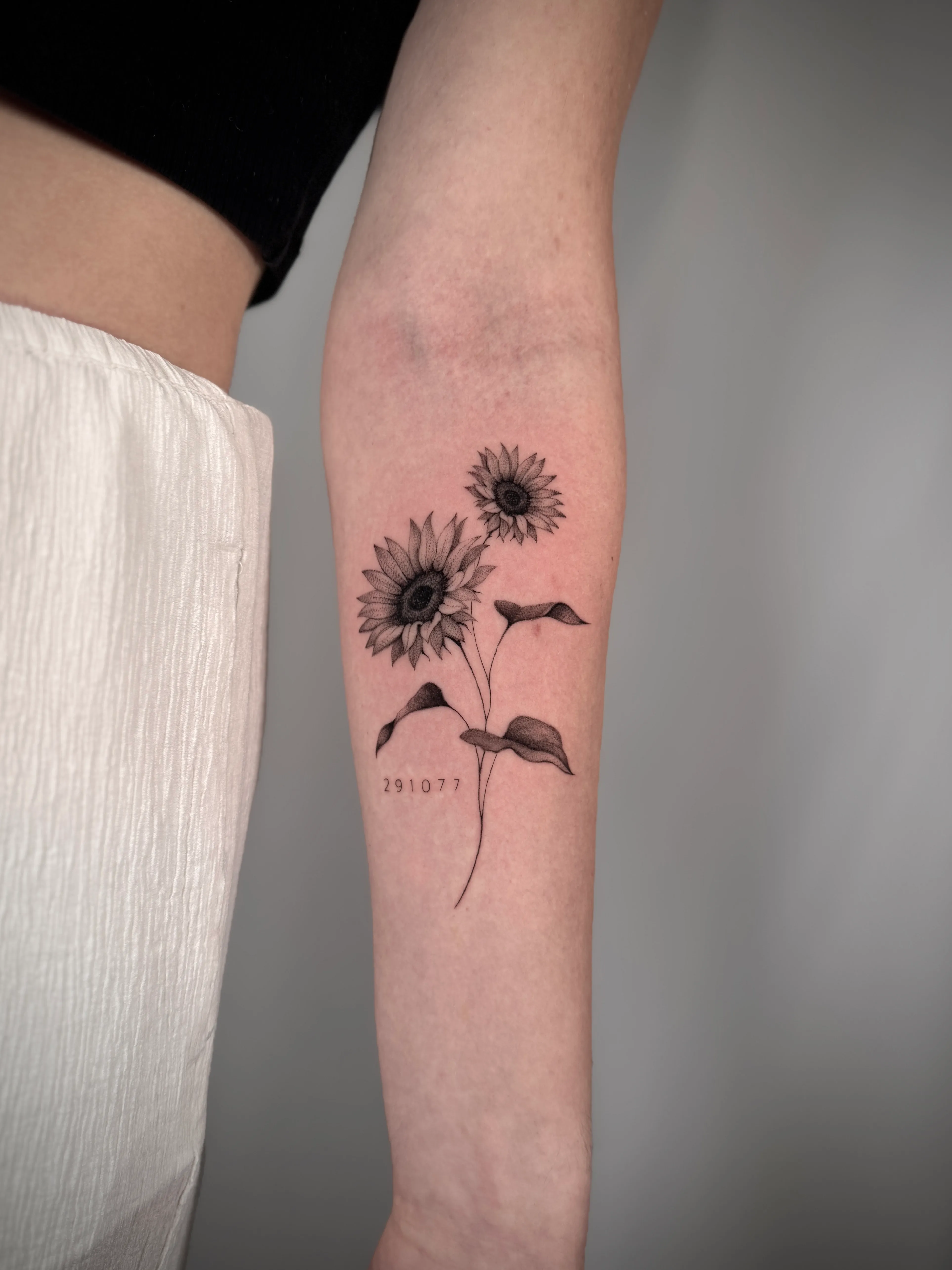 Tattoo auf Unterarm mit zwei detaillierten Sonnenblumen und der Zahlenfolge 291077 darunter.