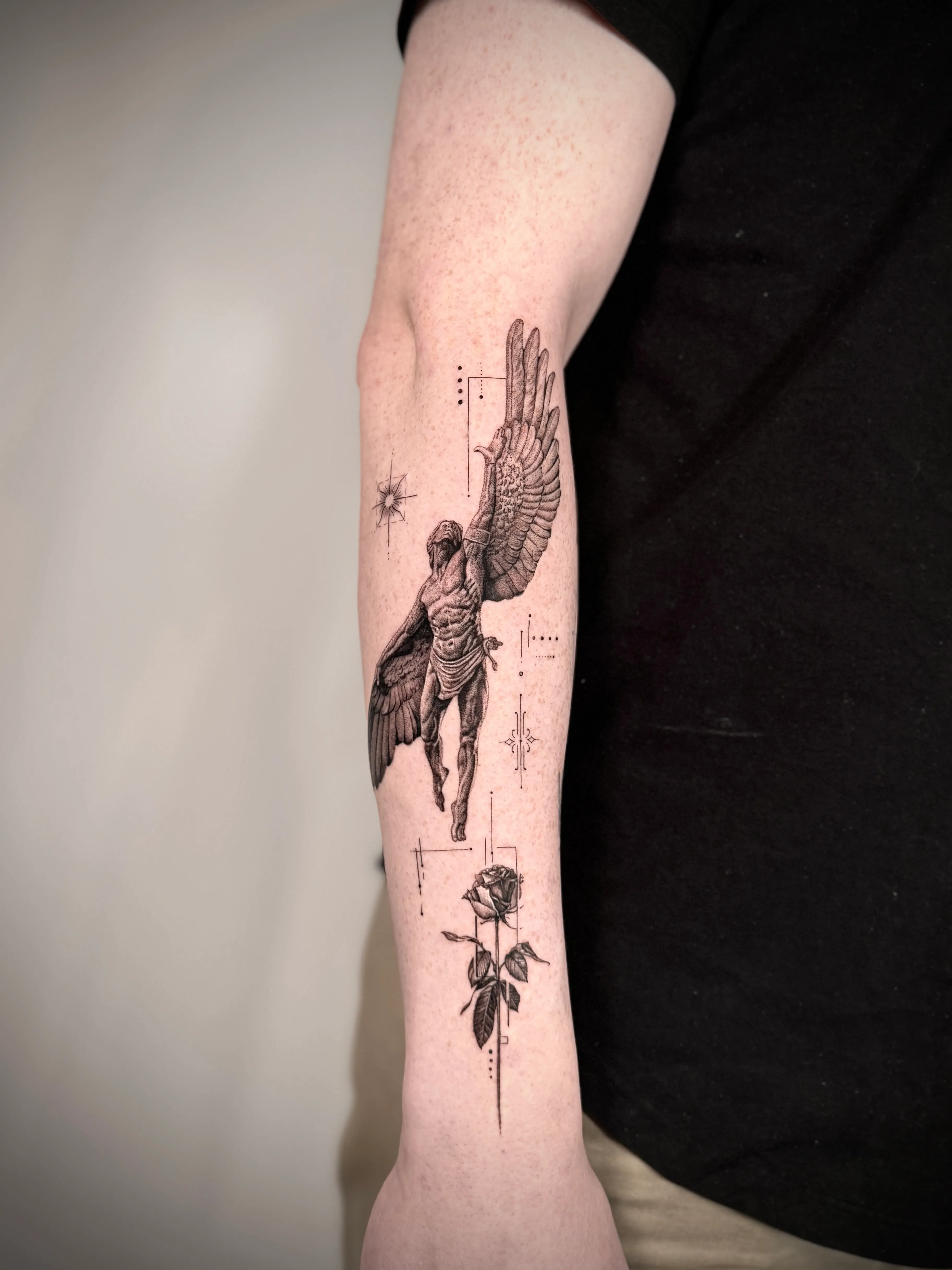Tattoo am Unterarm mit einer detaillierten männlichen Figur mit großen Flügeln, einer Rose und dekorativen Linien.