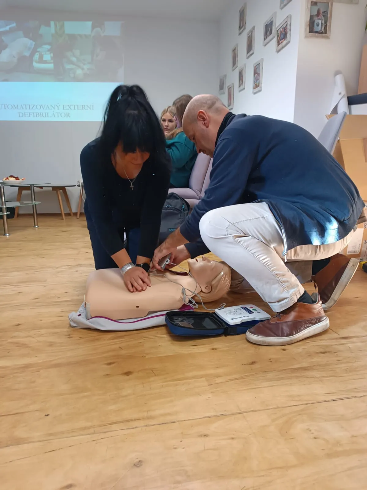 Two people practicing CPR on a training mannequin with an automated external defibrillator in a classroom setting.