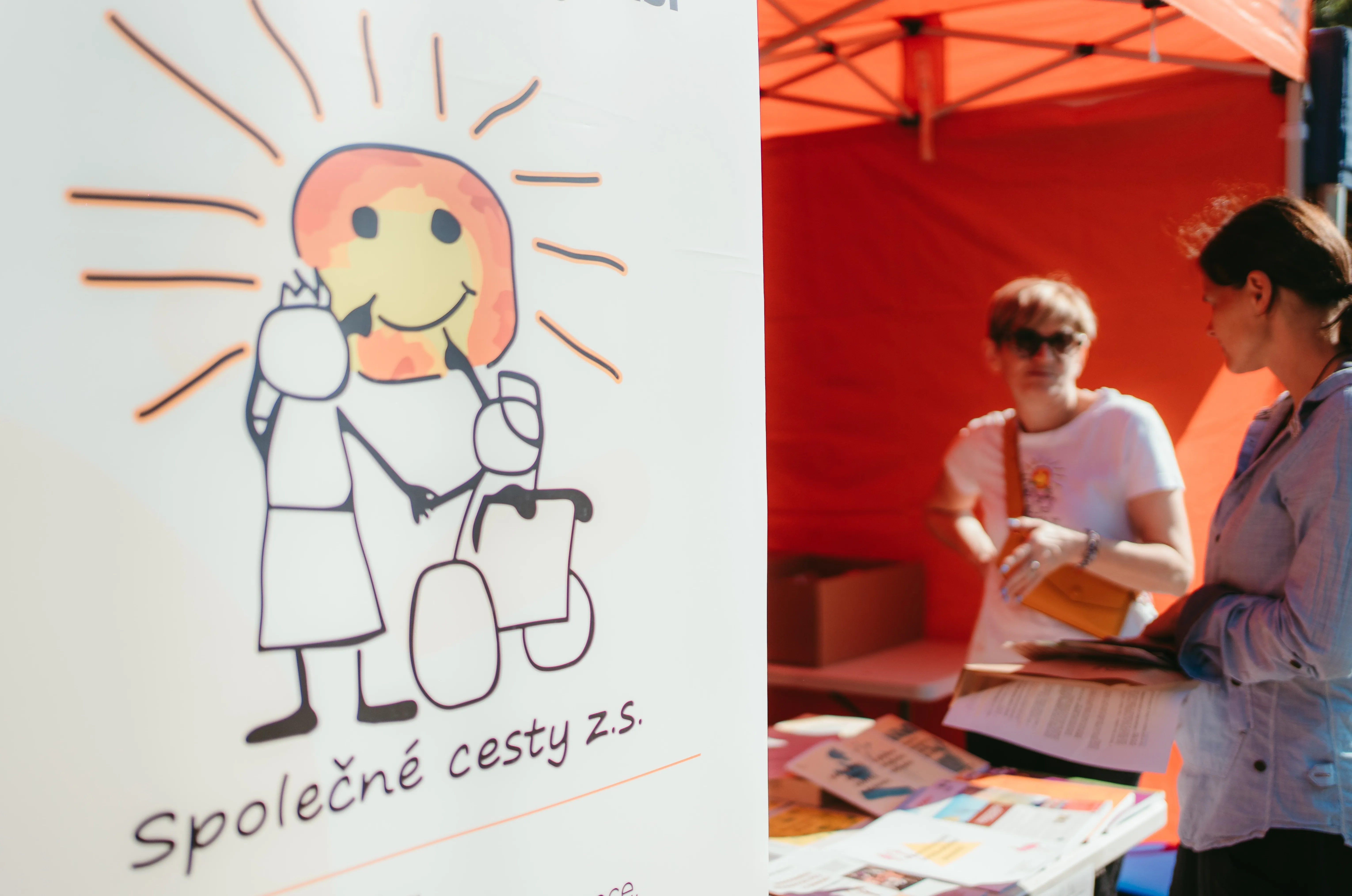 Two people talking beside a table with informational brochures under an orange canopy, with a white banner showing a sun-faced smiling figure pushing a wheelchair and the text 'Společné cesty z.s.'
