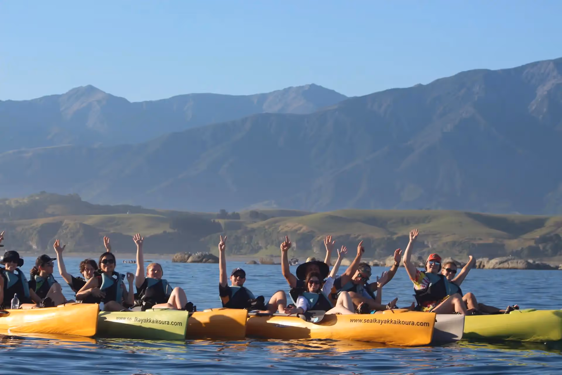 A group of people on kayaks with their arms in the air and the mountains behind them