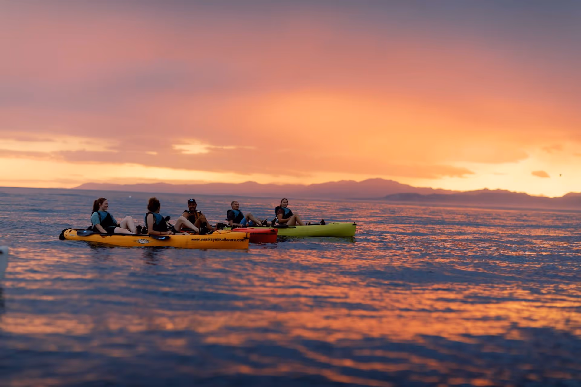Group of people on 4 pedal kayaks with the sun setting behind them on the water at Kaikōura