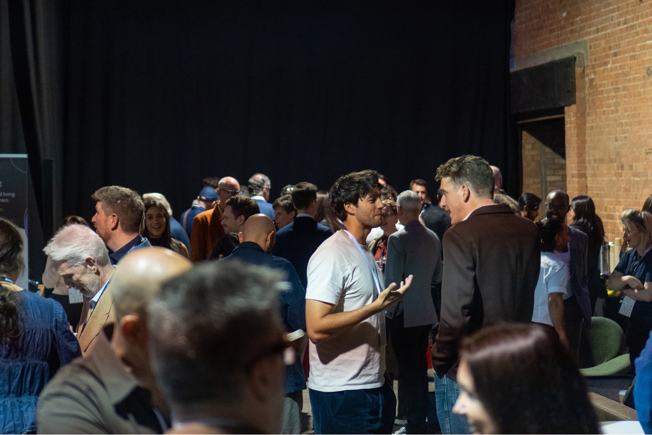 A crowded indoor networking during Coliving Conference with diverse people standing and engaging in conversations against a backdrop of black curtains and exposed brick walls.