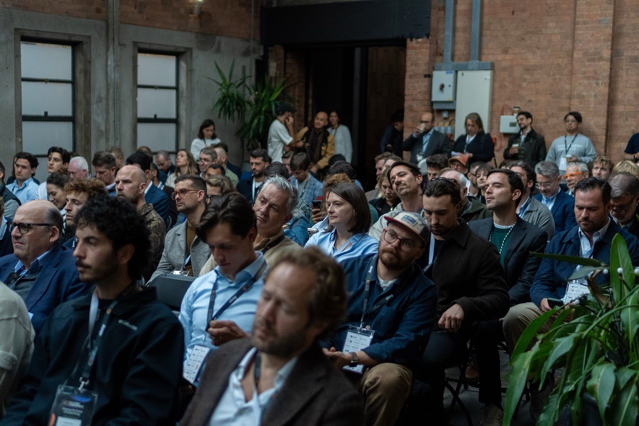Audience seated in rows inside a brick-walled venue of Coliving Conference, listening attentively at a conference or event, with some people wearing lanyards and badges.