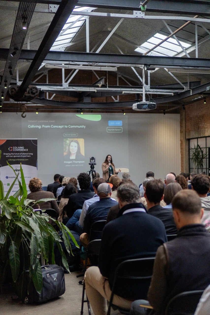 Indoor Coliving Conference setting with a woman speaking on stage to an audience seated in rows, presentation slide behind her titled 'Coliving: From Concept to Community' with speaker Imogen Thompson from Urban Land Institute, industrial ceiling with exposed beams and skylights, green plants on the left side.