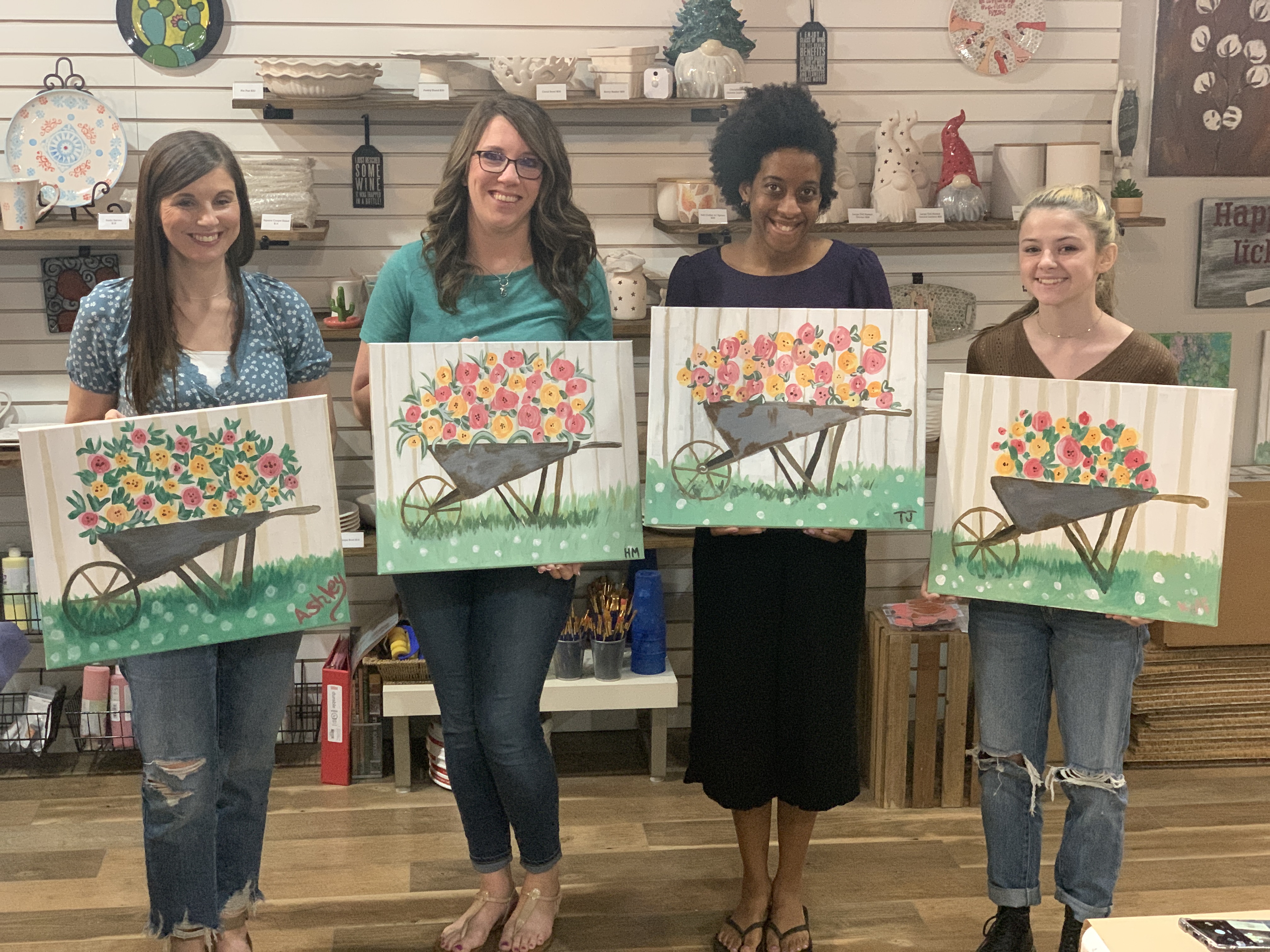 Four smiling women holding paintings of wheelbarrows with pink and yellow flowers in a studio.