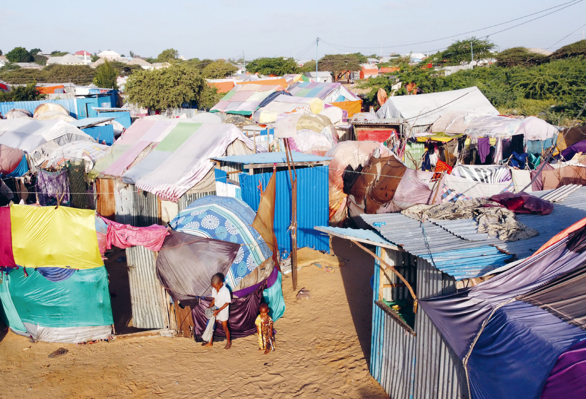 Gruppe afrikanischer Frauen sitzt im Freien auf dem Boden, umgeben von weißen Säcken mit Hilfsgütern; viele tragen bunte traditionelle Kleidung. Im Hintergrund hügelige Landschaft mit vereinzelten Bäumen.