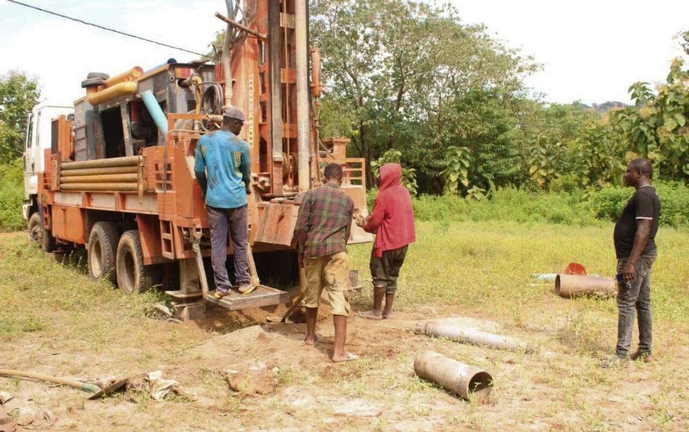 Gruppe afrikanischer Frauen sitzt im Freien auf dem Boden, umgeben von weißen Säcken mit Hilfsgütern; viele tragen bunte traditionelle Kleidung. Im Hintergrund hügelige Landschaft mit vereinzelten Bäumen.