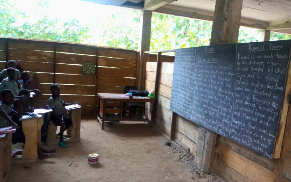 Einfaches Schulgebäude in kräftigem Türkisblau mit Blechdach in ländlicher Umgebung; zahlreiche afrikanische Schulkinder in Uniform spielen und sitzen auf dem sandigen Vorplatz unter Bäumen.