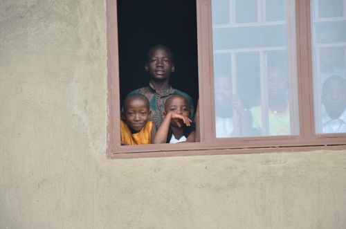 Zwei Kinder und ein Lehrer sehen aus dem Fenster eines unverputzten Neubaus.
