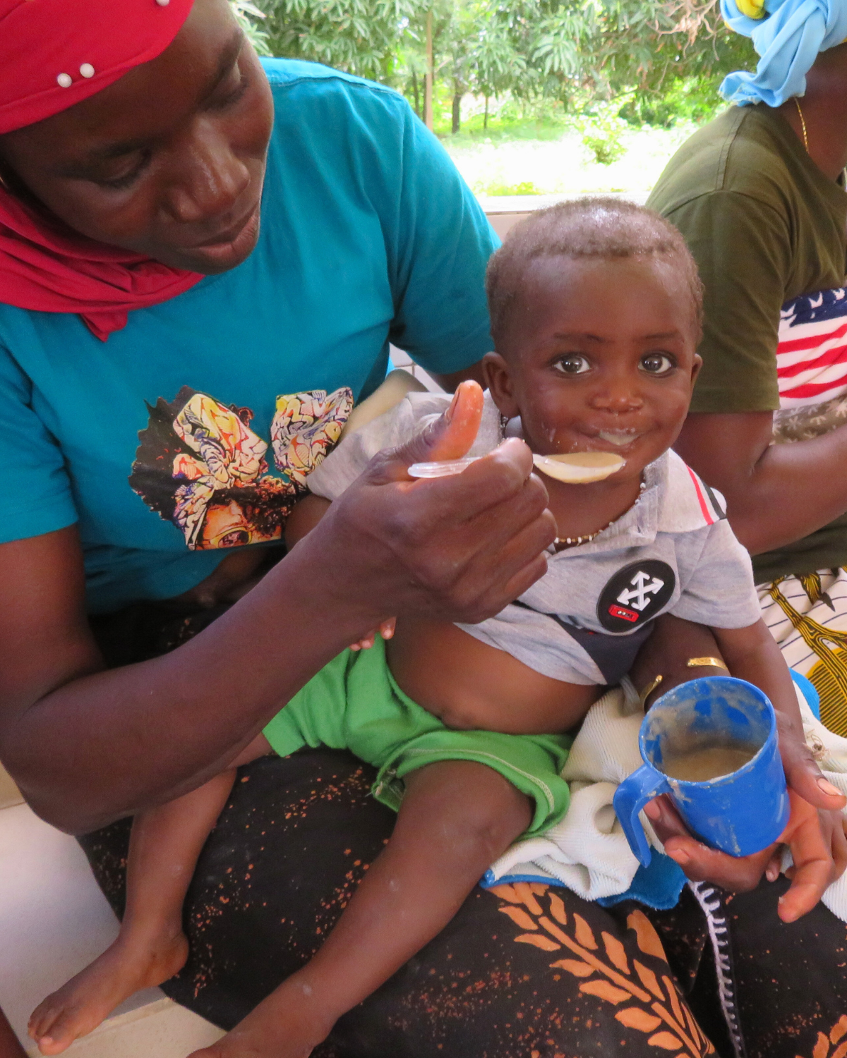 Eine Mutter in Burkina Faso füttert ihr Kleinkind mit nahrhaftem Brei aus einer blauen Tasse. Das Kind lächelt. Im Hintergrund sitzen weitere Frauen mit Babys. Sie alle sind Teil des Projekts «Nahrung für Kinder»-