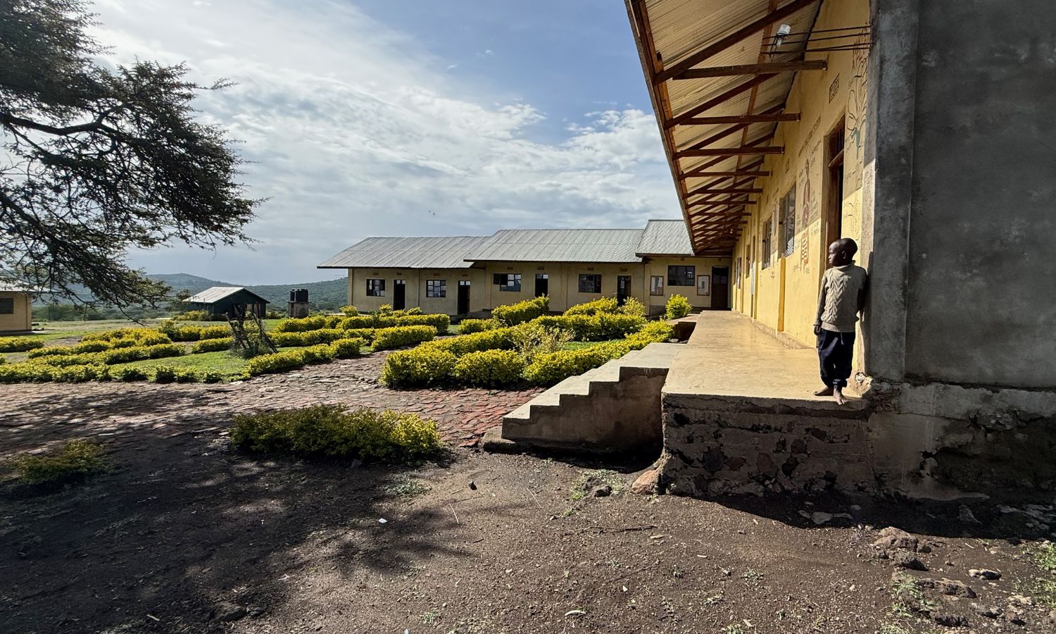 Ein Junge steht vor einem Schulgebäude in einer abgelegenen Region nahe der Serengeti in Tansania. Im Hintergrund ist die weite Savannenlandschaft zu sehen.