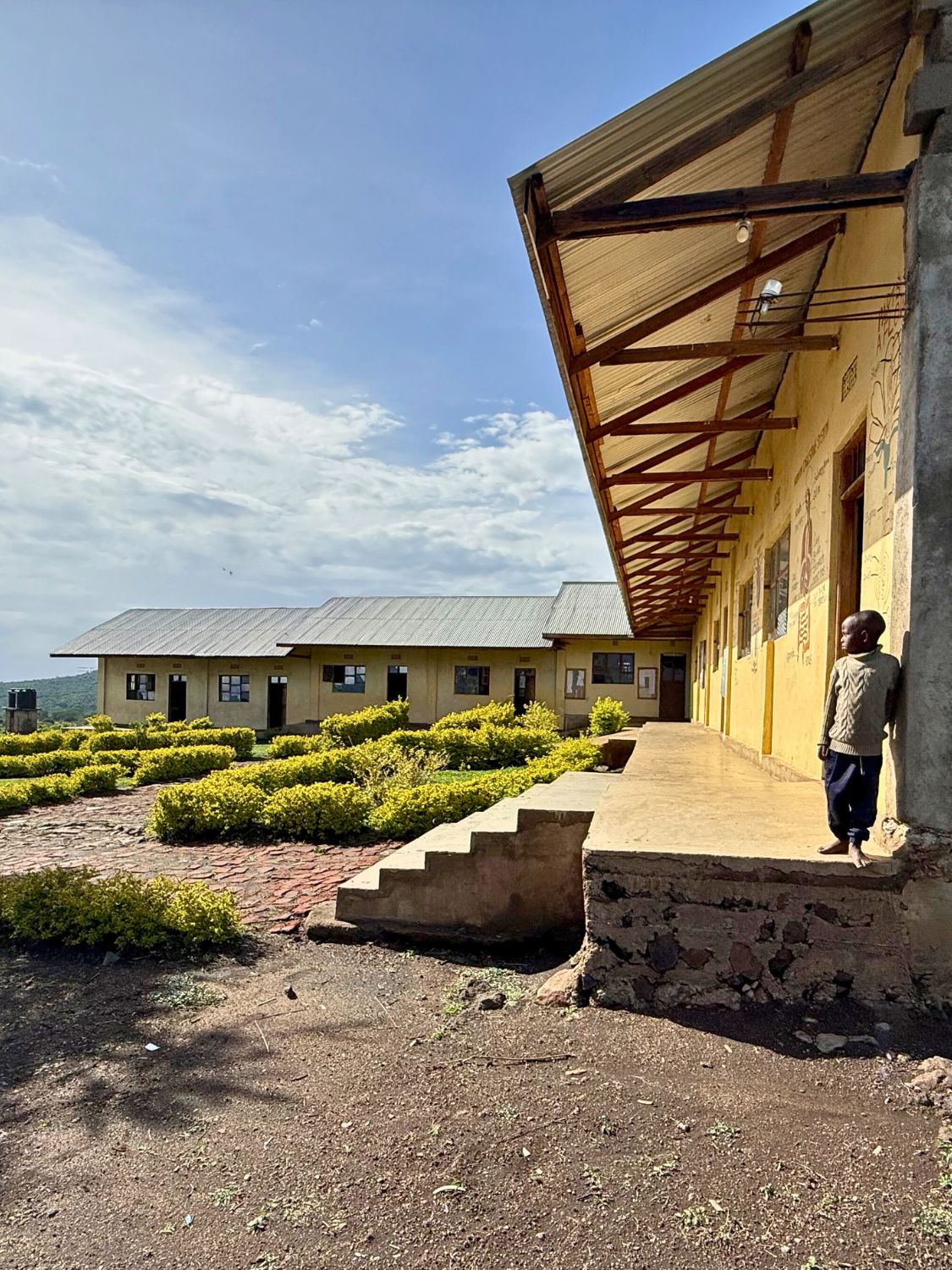 Ein Junge steht vor einem Schulgebäude in einer abgelegenen Region nahe der Serengeti in Tansania. Im Hintergrund ist die weite Savannenlandschaft zu sehen.