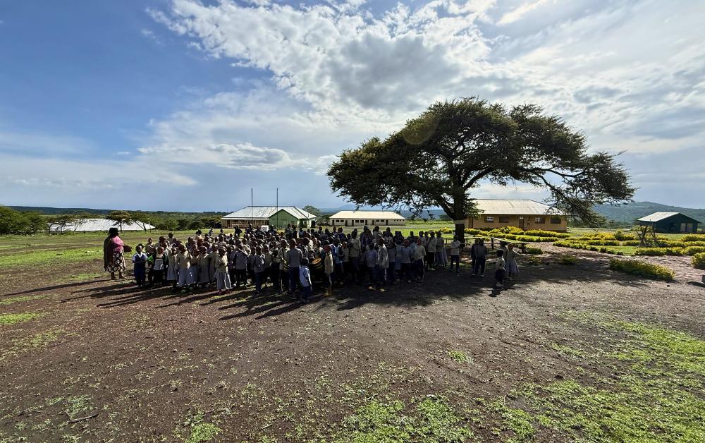 Eine Gruppe von Schulkindern steht unter einem großen Baum auf dem Schulgelände. Im Hintergrund sind mehrere Schulgebäude und eine hügelige Landschaft zu sehen.
