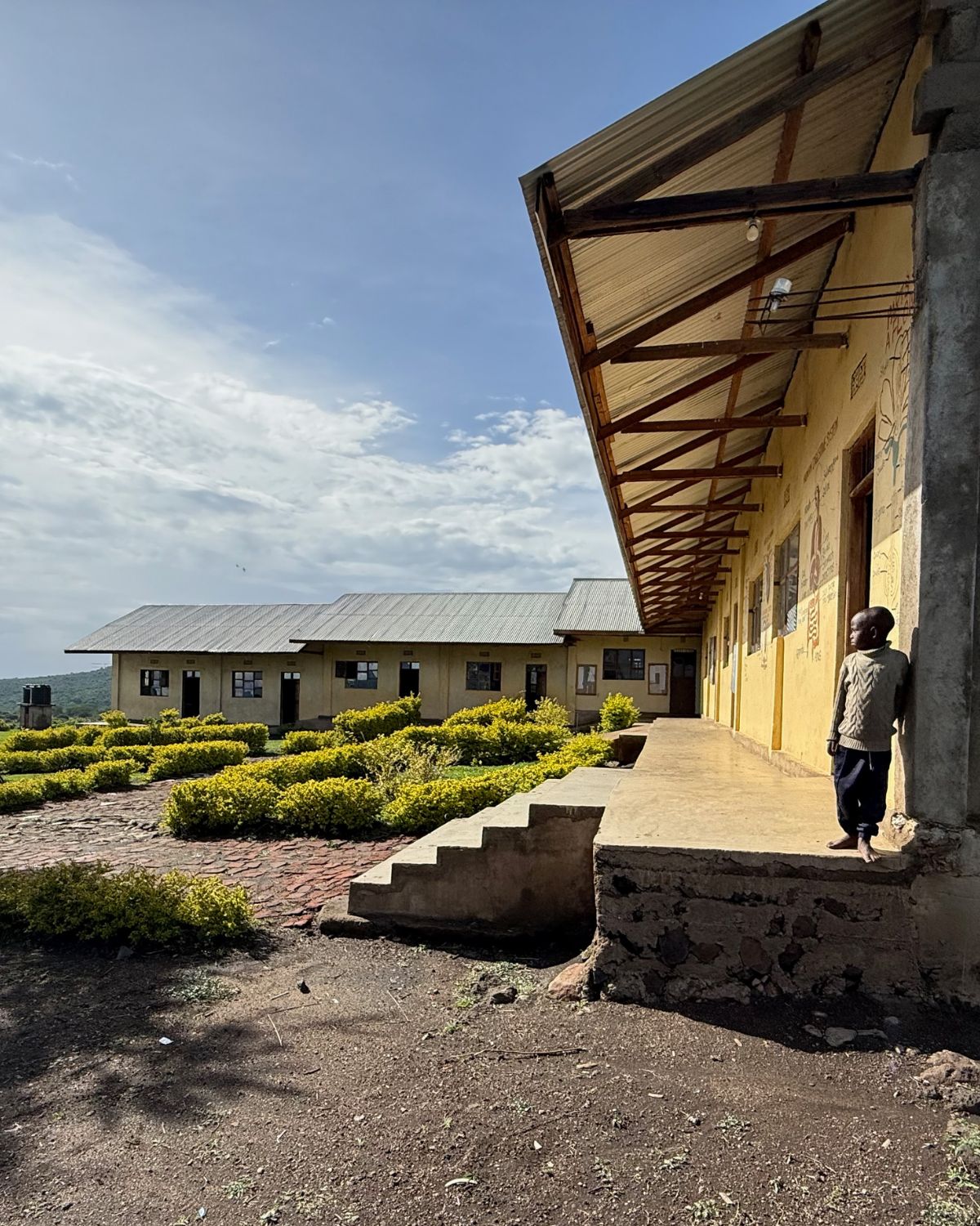 Ein Junge steht vor einem einfachen Schulgebäude der St Luke Pre-Primary School in Endulen, Tansania. Im Hintergrund sind die weite Savannenlandschaft und Hügel nahe der Serengeti zu sehen.