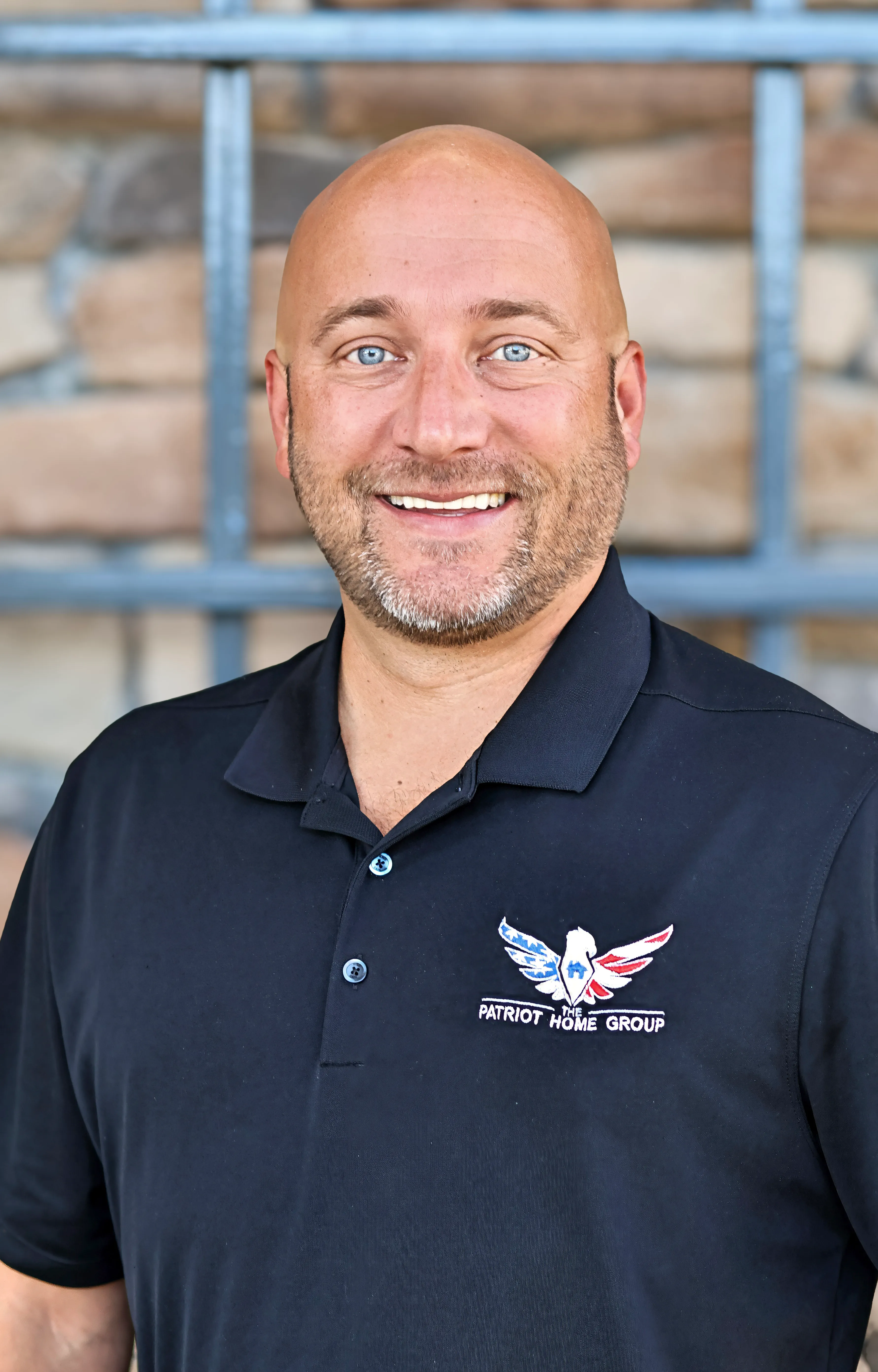 Smiling bald man with light beard wearing dark polo shirt with Patriot Home Group logo.