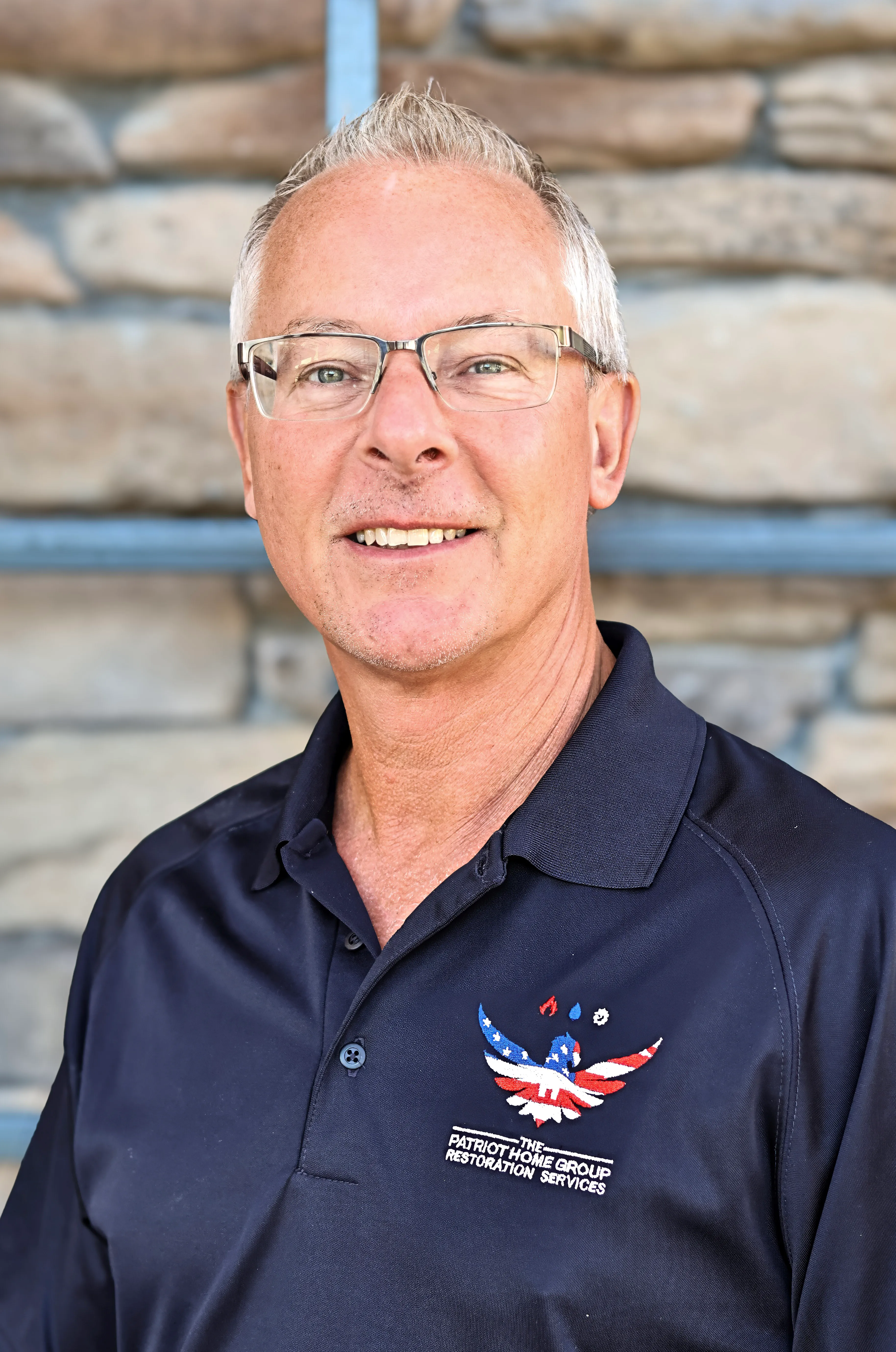 Smiling middle-aged man with glasses wearing a Patriot Home Group polo shirt, standing before a stone wall.