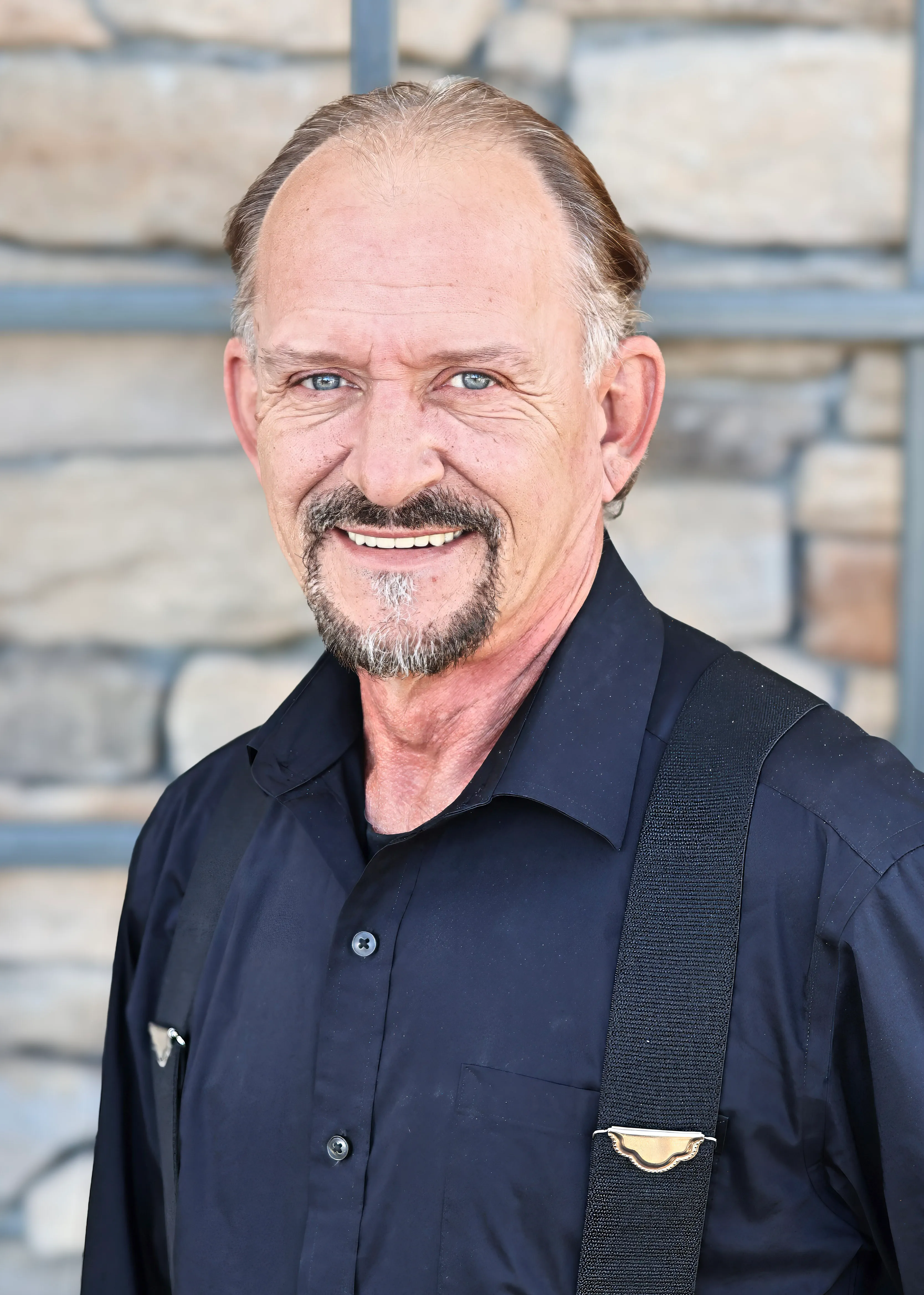 Smiling middle-aged man with goatee wearing a black shirt and suspenders in front of a stone wall.