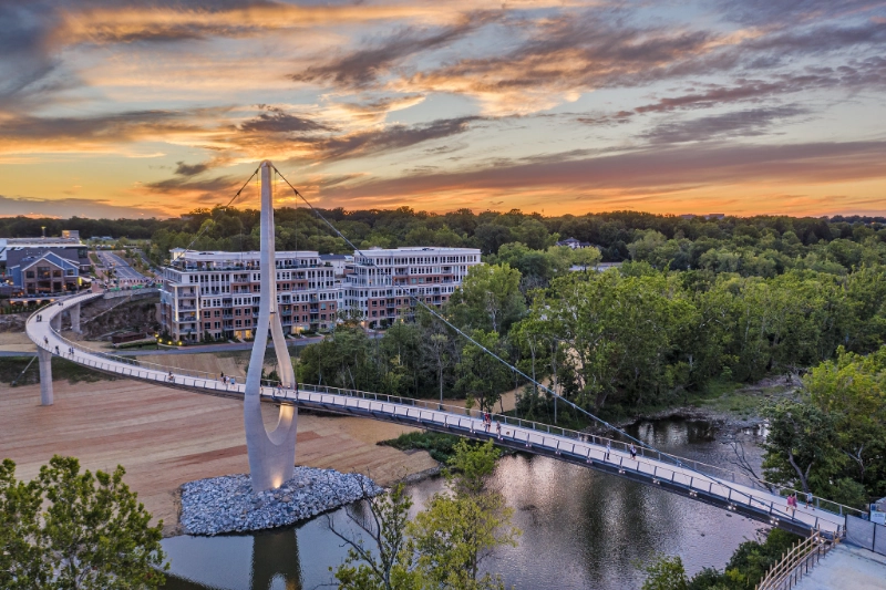 Suspension pedestrian bridge over a river near buildings at sunset with colorful sky and trees.