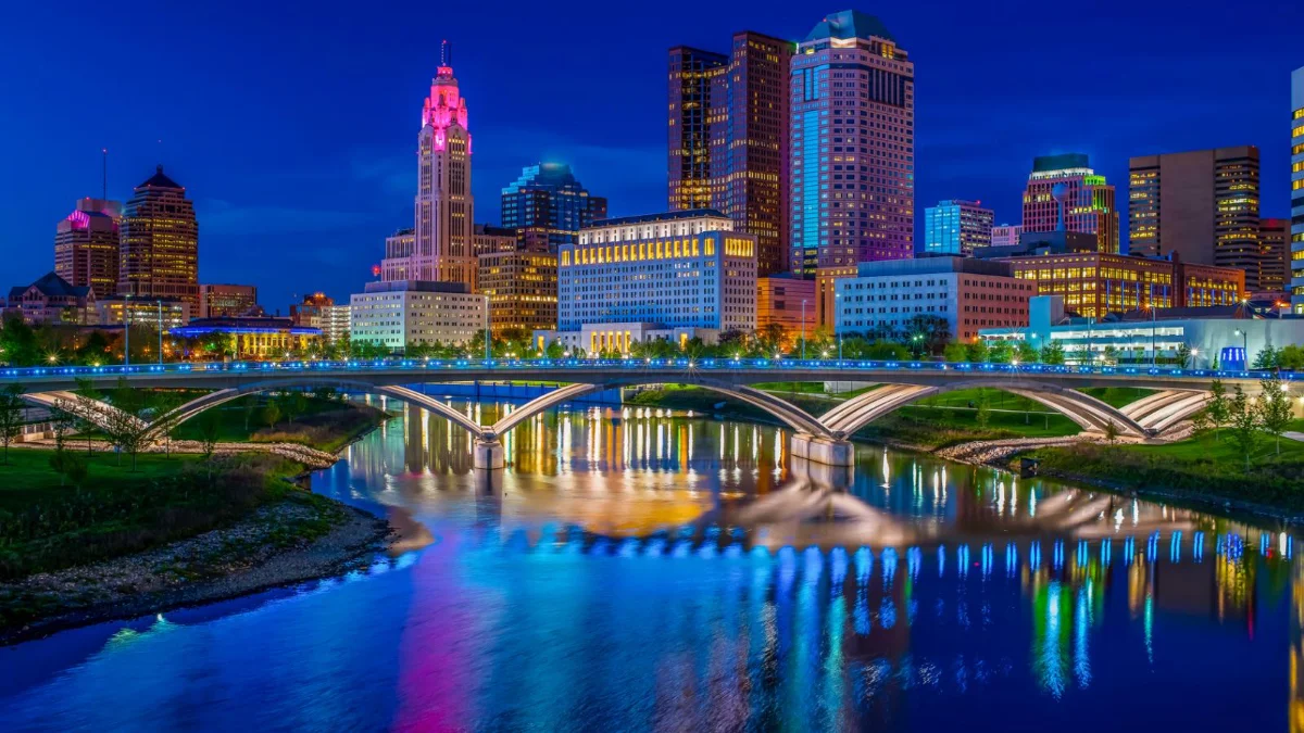 Night view of a city skyline with illuminated buildings and a bridge reflecting on the water.