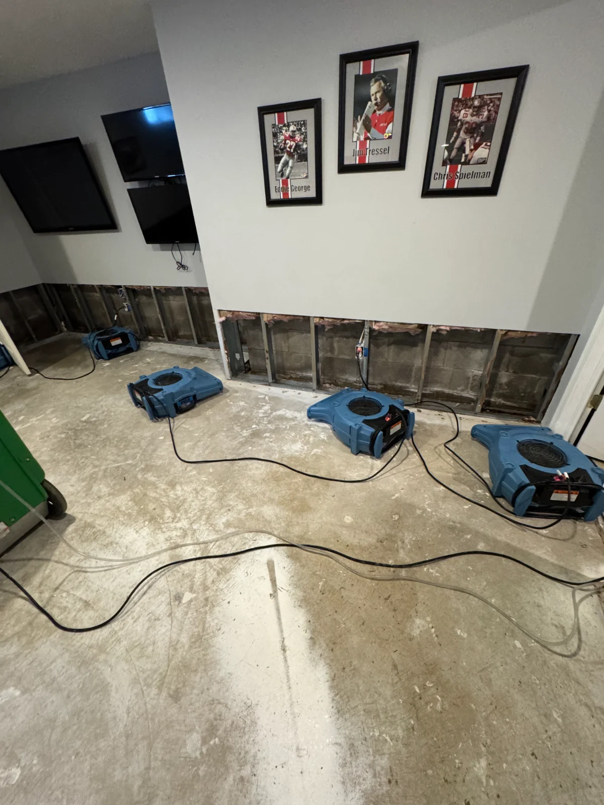 Room with lower drywall removed, three blue air dryers on floor, and three framed football photos on upper wall.