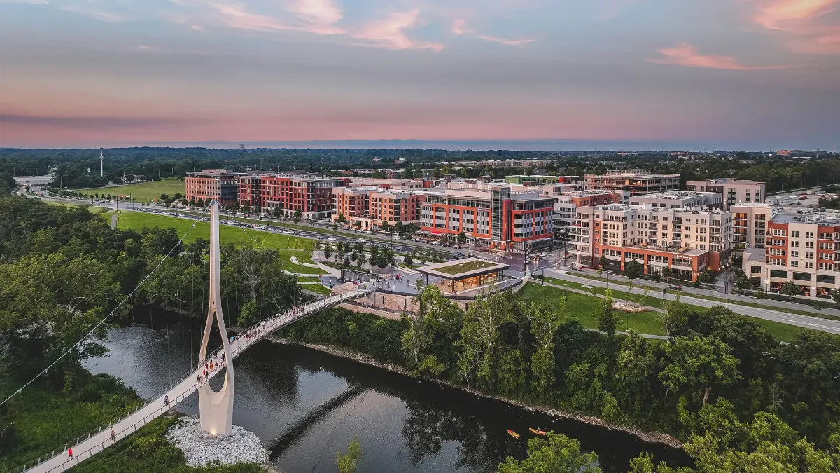 Suspension bridge over river with people, green park, and apartment buildings at sunset.