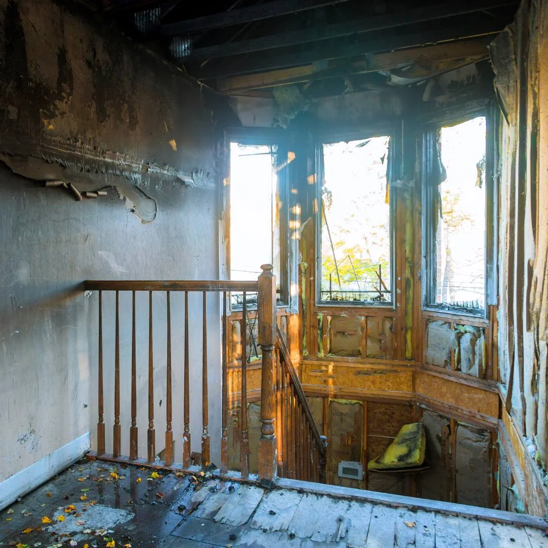 Fire-damaged interior with charred walls, broken windows, and a stained wooden railing by a staircase.