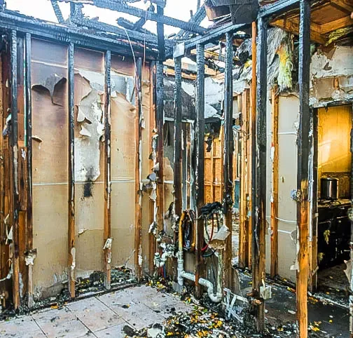 Interior of a house with extensive fire damage, showing charred wall studs and debris on floor.