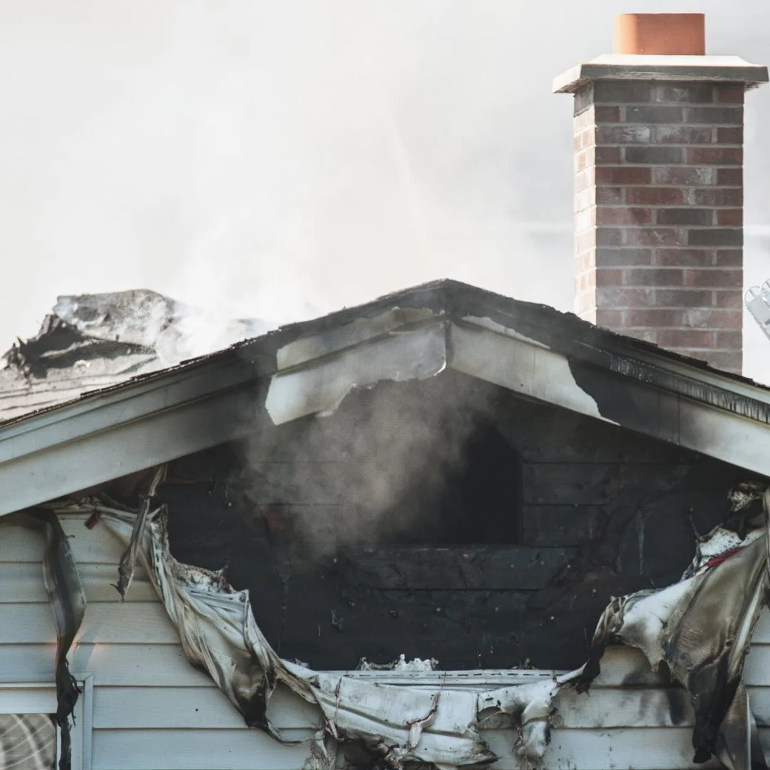 Front side of house with fire damage, smoke rising from burnt blackened siding near the roof.