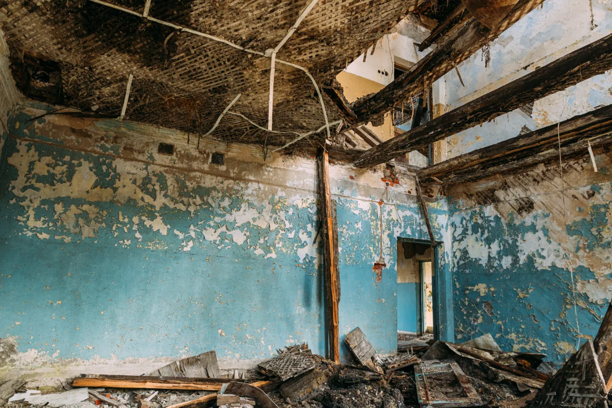 Interior of a room severely damaged by fire with charred wooden beams and peeling blue paint on walls.