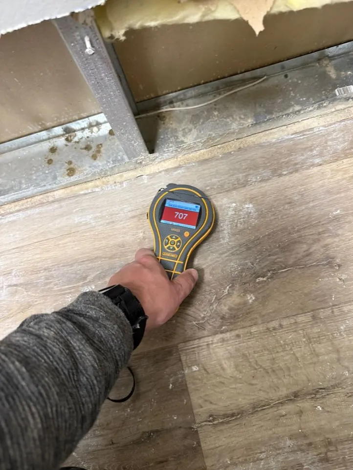Hand holding moisture meter showing 707 reading on floor near wall with metal studs and insulation.