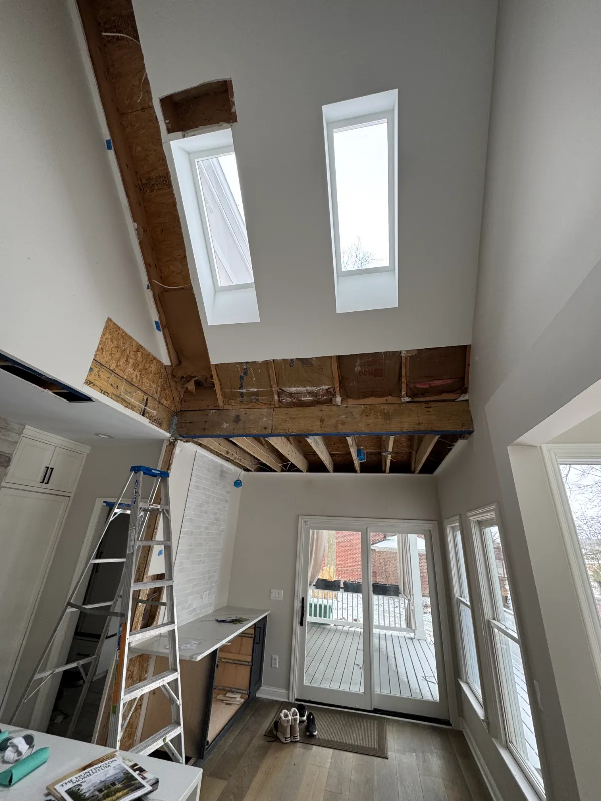 Interior of a home under renovation with exposed ceiling beams, skylights, ladder, and sliding glass door.