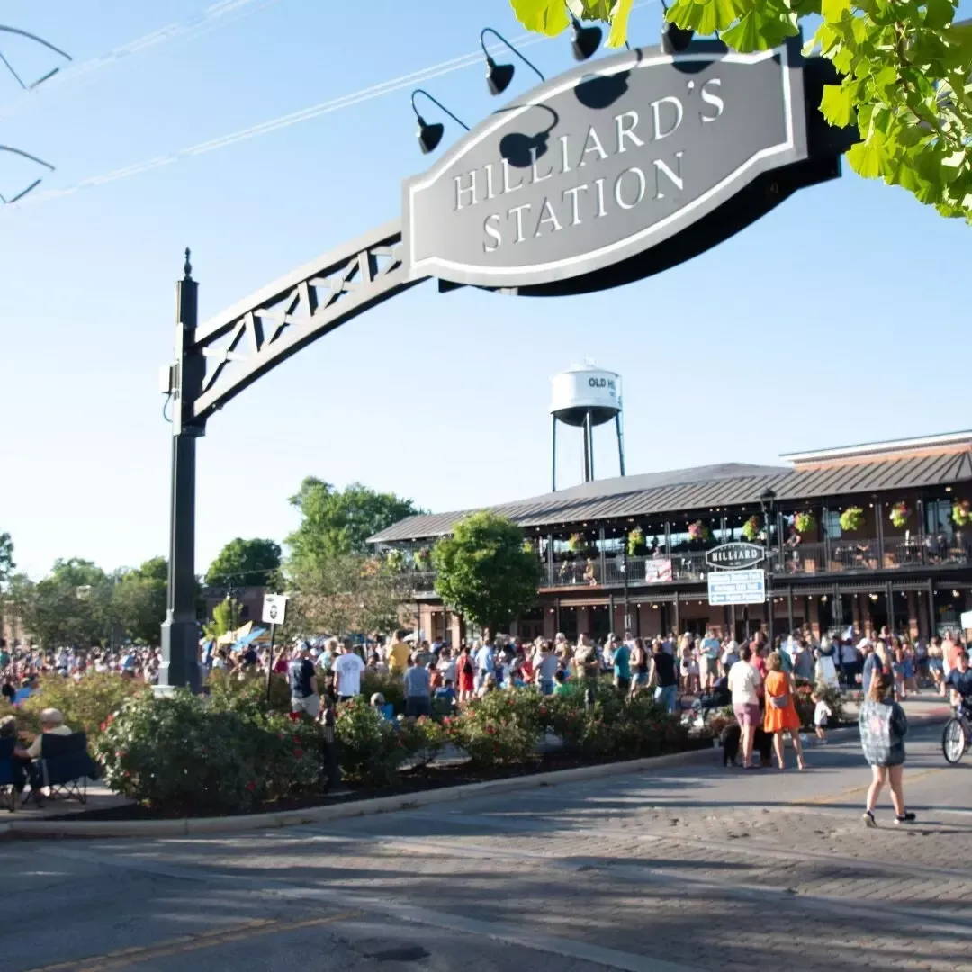 Crowd gathered under a large Hilliard's Station sign near a two-story building and water tower.