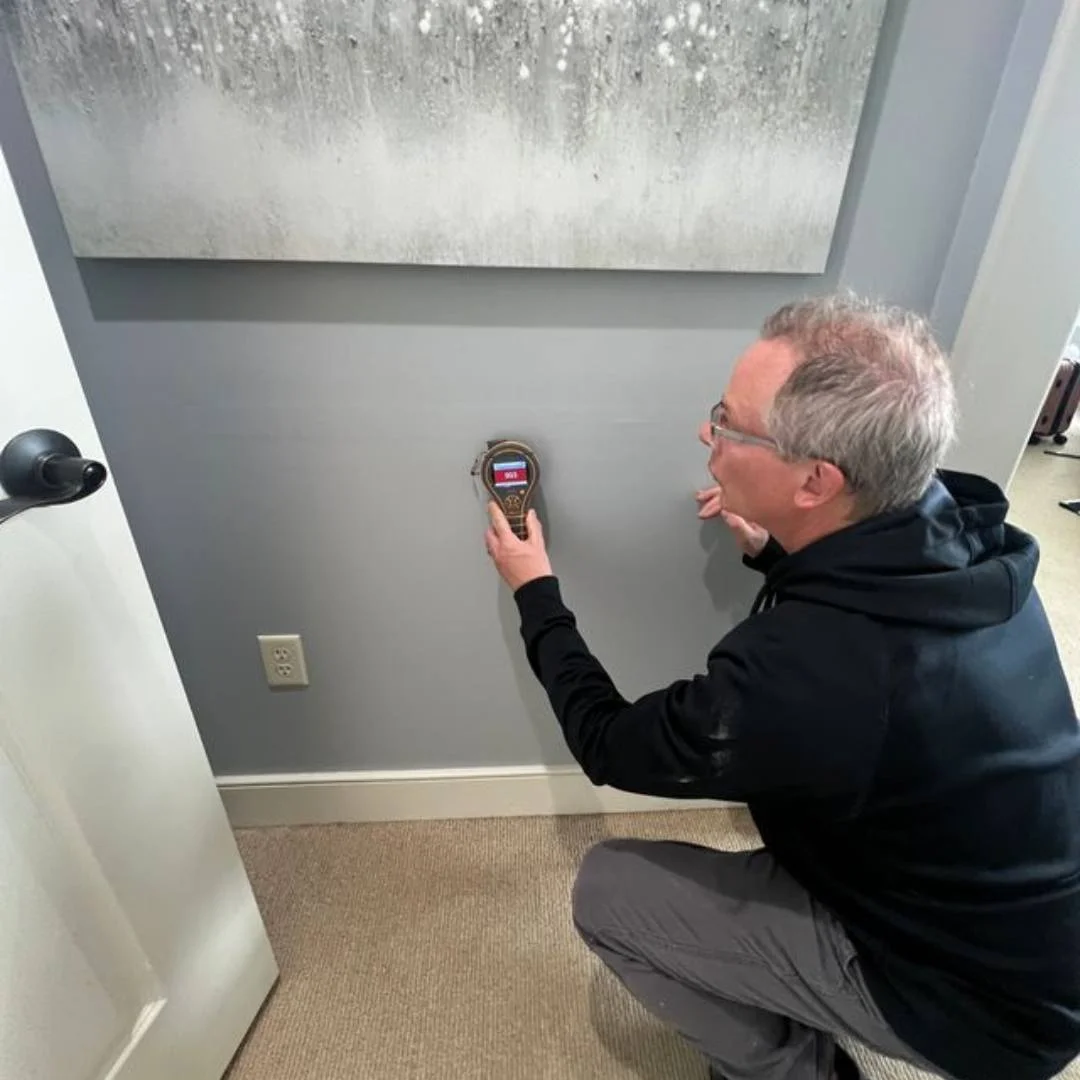 Man kneeling and using a handheld device to test or measure a gray wall in a room.