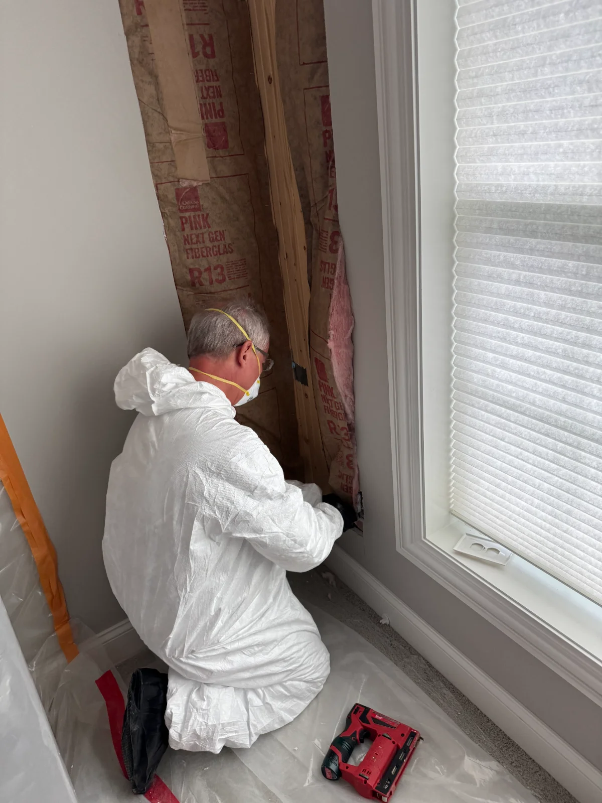 Worker in white protective suit and mask inspecting wall insulation near a window.