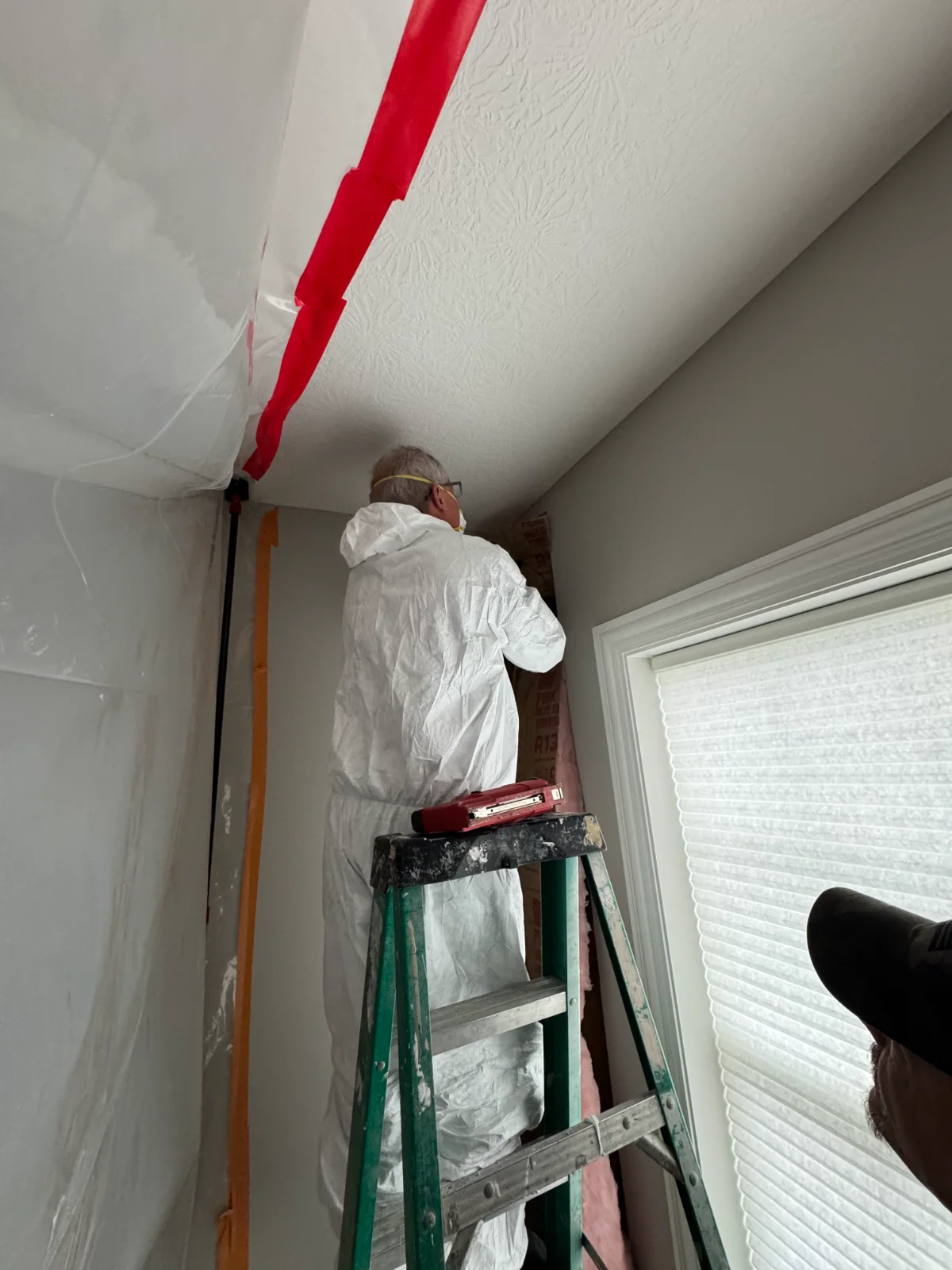 Person in white protective suit inspecting wall insulation on a green ladder near a curtained window.