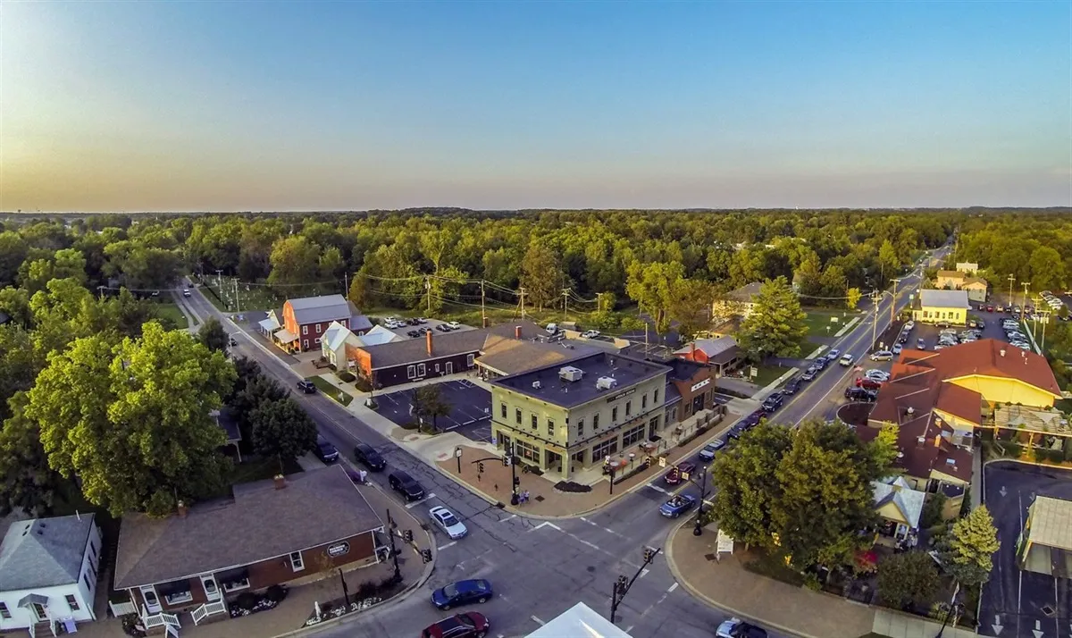 Aerial view of a town intersection with buildings, cars, trees, and a clear blue sky at dusk.