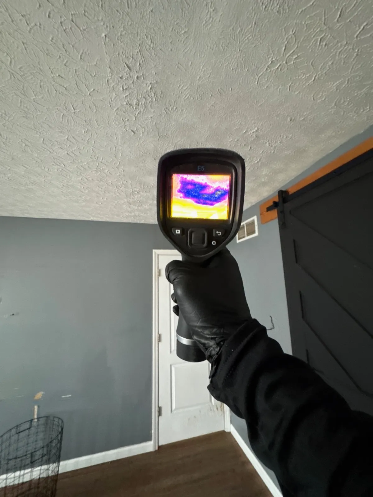 Hand in black glove holds thermal camera showing ceiling water damage with purple and yellow heat patterns.